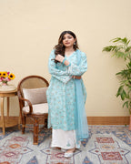 A woman posing in a blue and white floral print dress with a chiffon dupatta, standing in a room with a chair and a plant in the background.