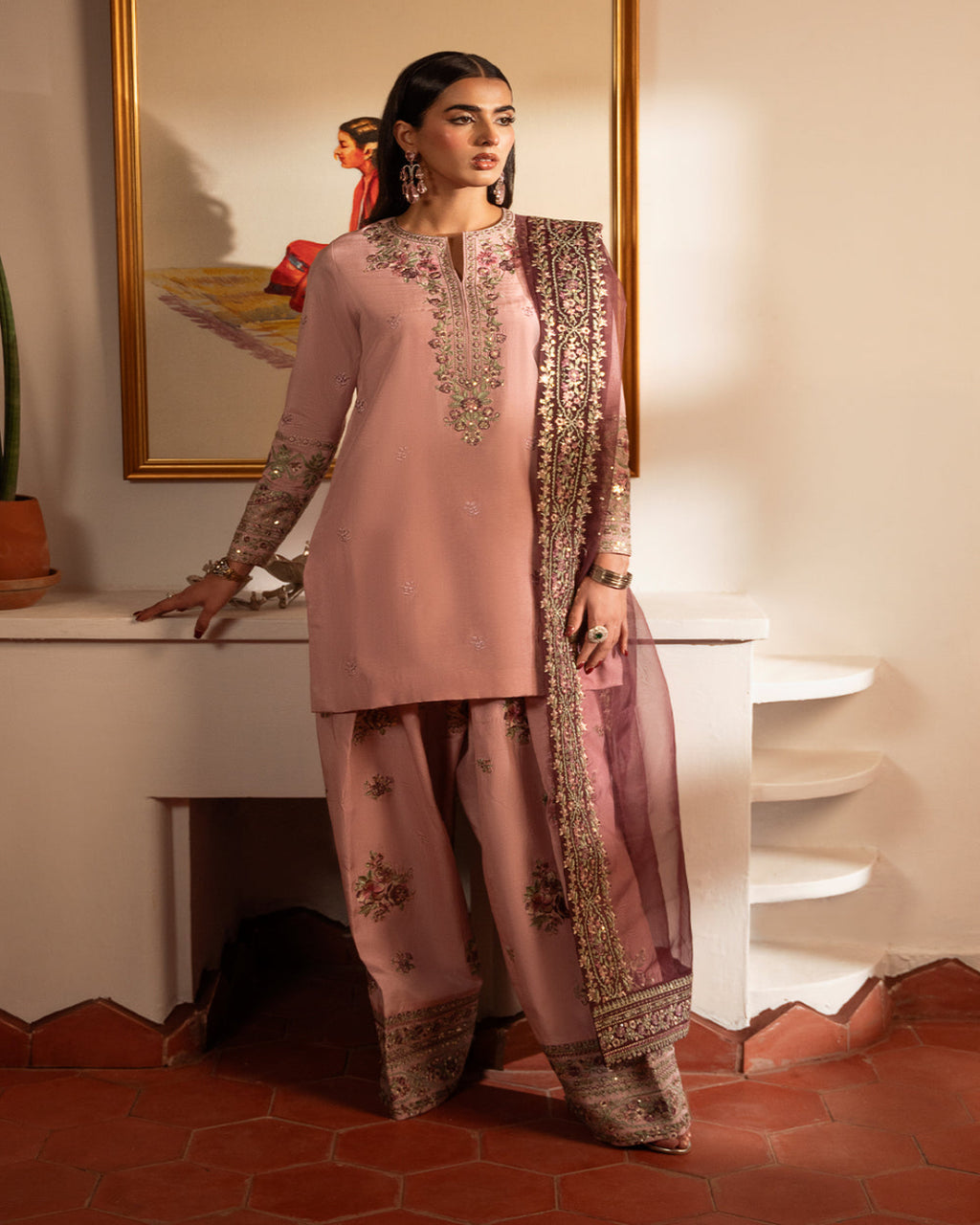 Woman in a traditional pink and gold outfit standing in a room with a mirror and decorative items.