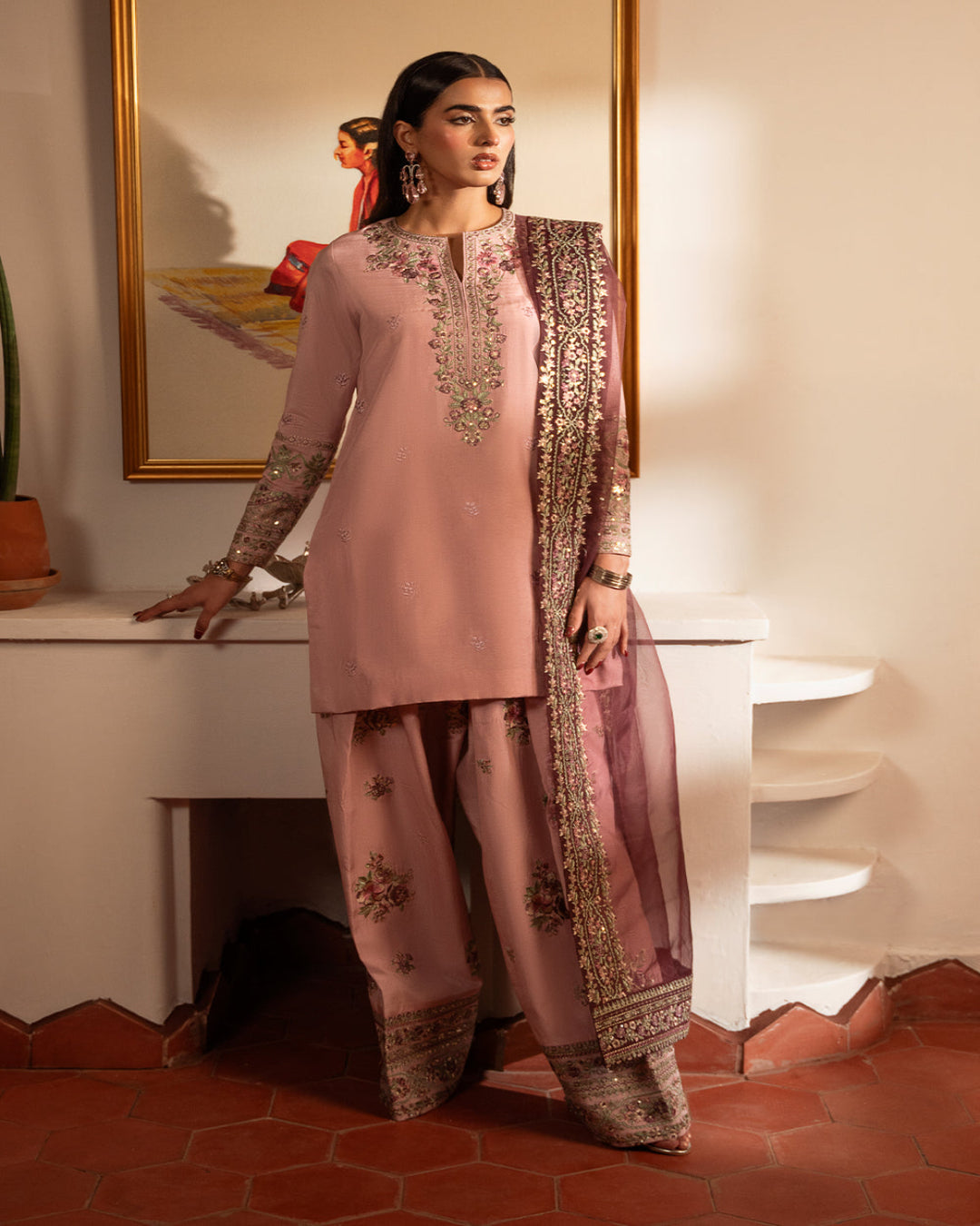 Woman in a traditional pink and gold outfit standing in a room with a mirror and decorative items.
